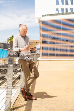 senior man reading newspaper standing on terrace near modern office building
