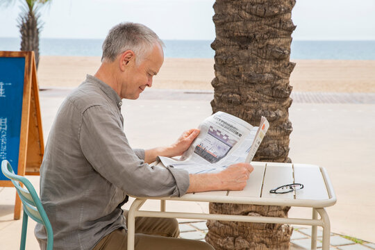 An elderly, gray-haired man is reading the news in a newspaper.