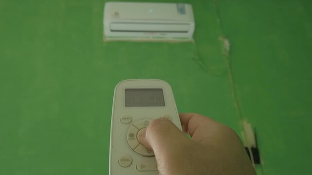 A person using an air conditioner remote control to turn on an indoor air conditioning unit. The scene represents home comfort, temperature control, cooling technology, and everyday household activiti
