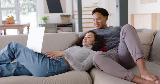 African American couple on sofa at home, woman scrolling laptop showing post, man laughing