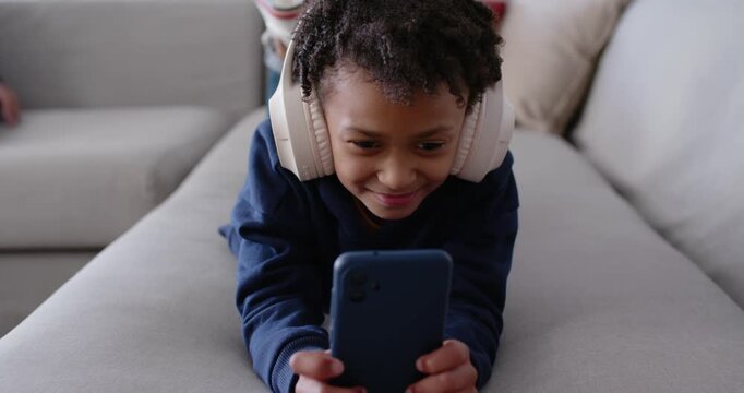School-age girl lying on gray sofa holding blue phone, wearing cream headphones