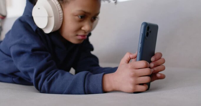 African American girl child tapping blue phone lying on sofa wearing headphones and navy sweatshirt