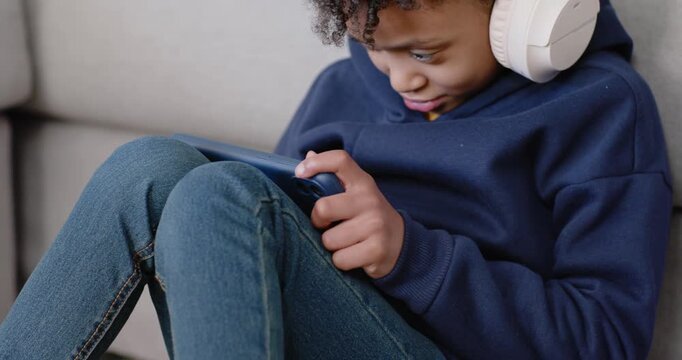 African American teenage girl holding blue tablet, wearing white headphones, adjusting grip on sofa