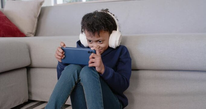 School-age African American girl at home in white headphones holding blue phone, reacting to game