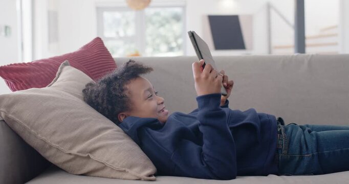 School-age African American girl lying on grey couch raising tablet and reacting to screen content