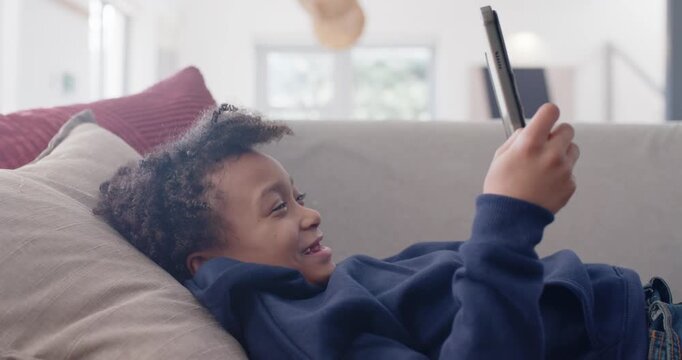 African American child lying on sofa in navy hoodie holding tablet, reacting to screen content