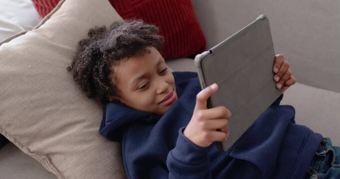 African American schoolboy raising tablet, tapping screen, reacting to content on couch in hoodie