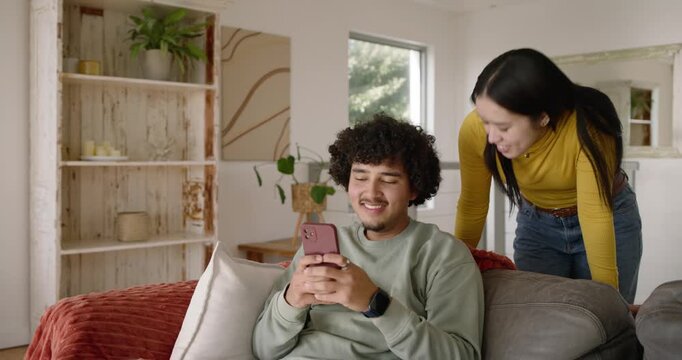 African American couple on couch, woman entering leaning to show pink phone, man scrolling reacting