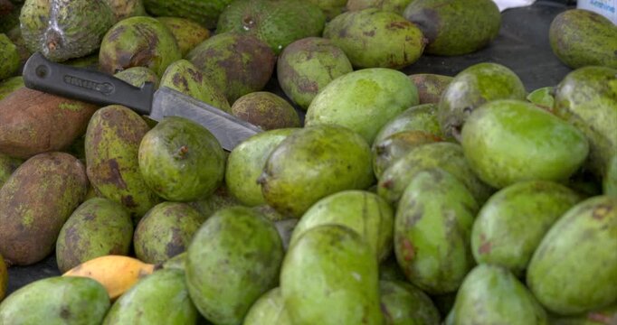 Pile of green mangoes rests beside a large cutting knife on a market counter with worn containers soursop and other tropical produce layered through the background in a static close composition