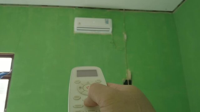 A person using an air conditioner remote control to turn on an indoor air conditioning unit. The scene represents home comfort, temperature control, cooling technology, and everyday household activiti