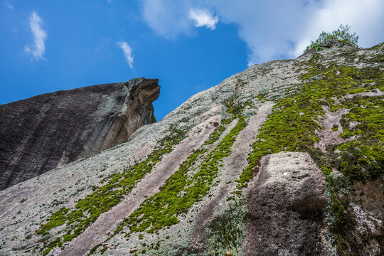 A massive mossy granite cliff with a dramatic rocky overhang rises steeply towards blue skies and scattered clouds.