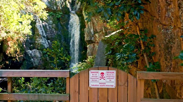A wooden fence and barrier with a red warning sign with a skull and cross bones, preventing hikers from going further along a path and risking their safety near a waterfall in South Africa