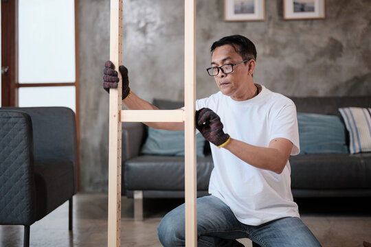 Asian mature man self-assembling flat-pack furniture with DIY tools, checking accuracy and strength of wooden parts, home improvement, craft installation, hobby workshop, handyman active lifestyle.