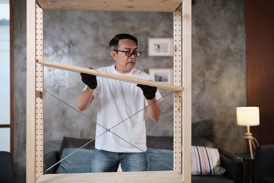 Asian mature man in gloves determined to assemble furniture with DIY tools, installing strong wooden shelf, home improvement, craft making, hobby carpentry workshop, expert handyman, active lifestyle.