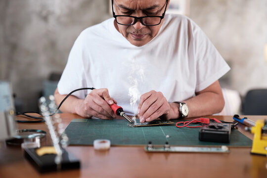 Mature Asian man repairs home electrical appliance, disassembling and soldering circuit board, focusing on precision detail, DIY fix tools, electronics component workshop hobby, expert handyman.