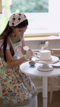 Young Asian cake decorator smoothing buttercream frosting on white cake with spatula in bright home bakery studio
