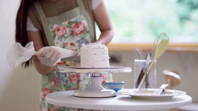 Young Asian woman baker decorating pink cake with spatula on stand in bright home bakery kitchen