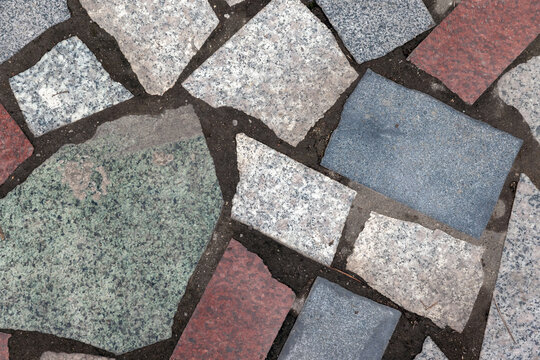 Closeup of a multicolored stone mosaic pavement showing irregular granite and flagstone pieces set in dark grout, highlighting texture, pattern and weathered outdoor hardscape detail