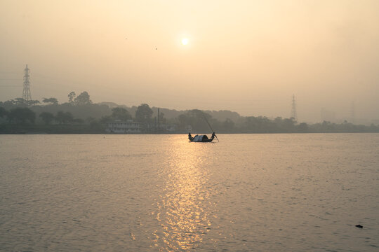 A small boat with a fisherman rowing on a calm river during a golden sunset with hazy skies and distant trees in the background