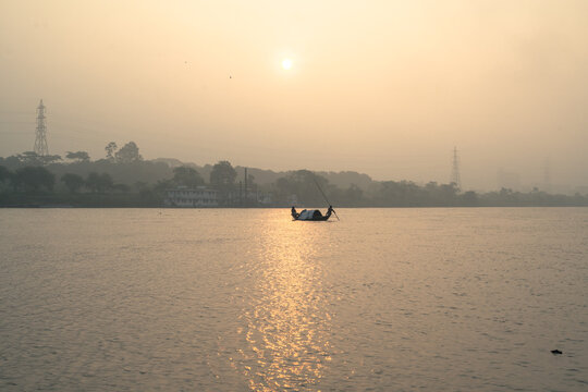 A small wooden boat with two people rowing on a calm river during a hazy golden sunset in india