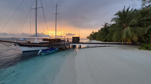 A steady wide view shows a two-masted wooden yacht and small motorboat moored at a palm-lined jetty beside a white sand beach as sunset glows beneath heavy clouds in the Maldives.