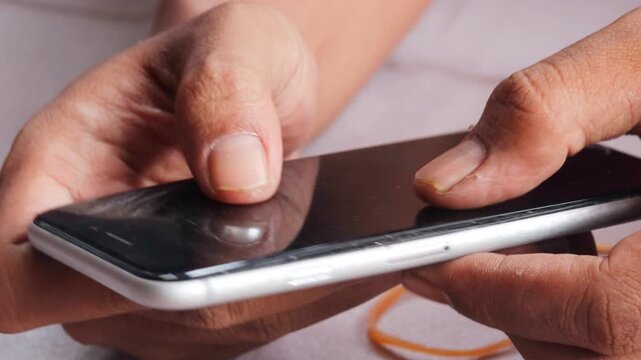 Close-up of Asian man's hands securing a slightly lifted smartphone screen with a rubber band as a temporary repair. Phone maintenance, financial hardship, and extending device use.