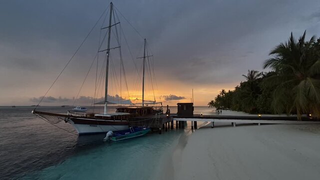 Wide coastal view of a wooden sailing yacht moored beside a palm-lined island jetty, with a golden sunset over turquoise water in the Maldives.