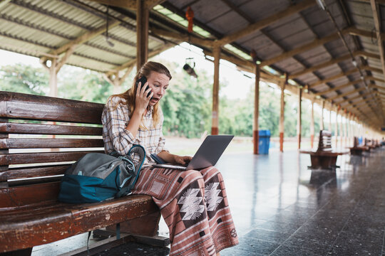 Woman traveler feeling shock while talking on phone and using laptop at train station