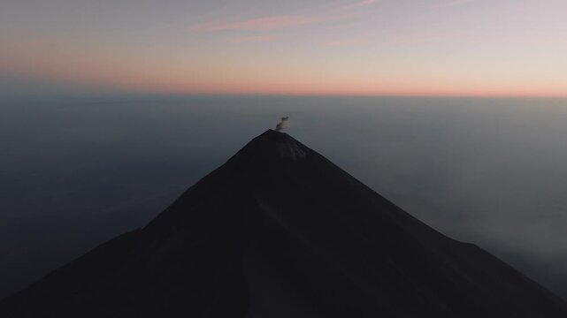 Drone shot captures Volc&aacute;n de Fuego erupting at sunrise with plumes of smoke, dark volcanic peak, glowing sky, and rolling low clouds, shot from Acatenango.