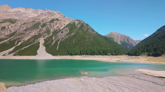 Turquoise mountain reservoir Lake of Livigno in Italy framed by steep rocky slopes, low water, clear blue summer sky. Pan right motion.