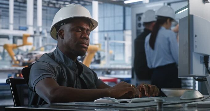 African American Engineer in Hard Hat Types Computer Workstation in Smart Factory Control Room. Background Colleagues Check Server Racks While Robotic Arms Assemble Vehicles. Concept of Robotization.