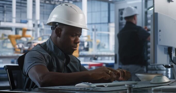 Inside Automated Plant Control Office, African American Specialist Types on PC Keyboard to Manage Production Algorithms. Network Technicians Service Server Hardware in Background. Industry 4.0.