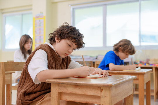 Focused schoolboy with curly hair writing with pencil in notebook at desk in bright elementary classroom with multiethnic classmates