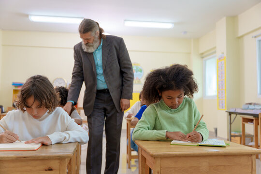 Senior male teacher observes diverse elementary students writing in notebooks at wooden desks in a bright classroom focused young schoolgirl in green sweater