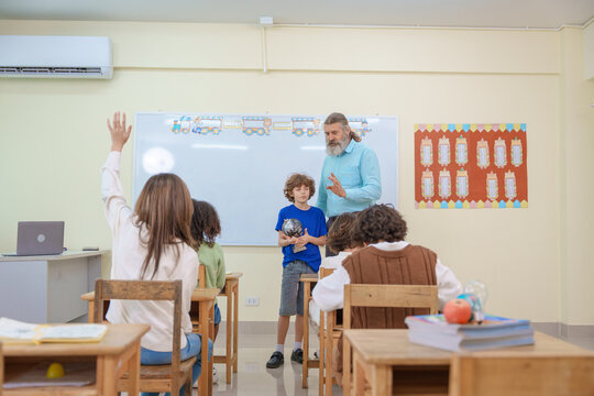 A senior teacher and a young student holding a globe present a geography lesson to diverse children in a bright classroom with a student raising her hand