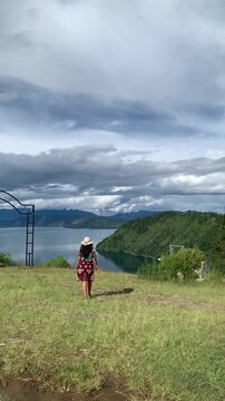 Vertical wide shot of a woman tourist walking across a grassy hill toward the beautiful vista of Danau Toba in Sumatera Utara, Indonesia, ideal for vacation, eco-tourism, and travel advertising.