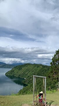 Vertical wide shot of a tourist woman swinging on a hilltop while admiring the scenic view of Lake Toba in Sumatera Utara, Indonesia, perfect for travel, freedom, and vacation concept projects.