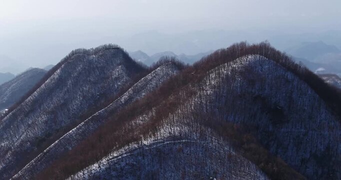 Aerial drone view of snow-dusted Qinling Mountains ridges in winter, bare deciduous forest on steep slopes with winding mountain road