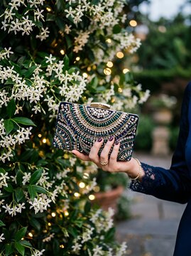 Woman holding ornate beaded clutch bag in front of blooming jasmine flowers and fairy lights.