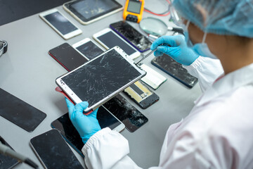 Technician wearing gloves repairs a cracked tablet with tools at a workbench covered with damaged smart phones and mobile devices. © NanSan