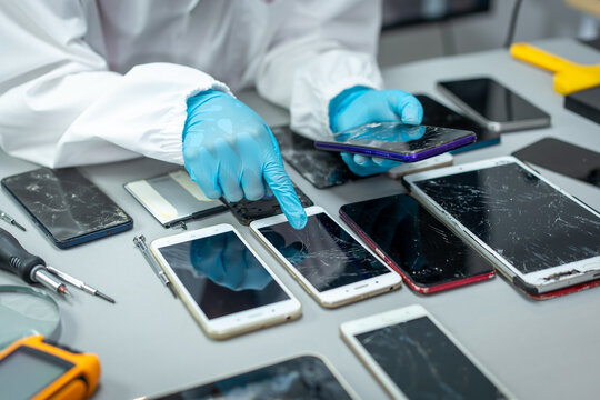 Technician in blue gloves examines damaged smart phones on a workbench, sorting cracked screens and devices during electronic repair.
