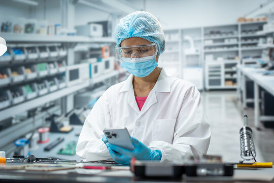 Female technician in protective clothing uses a smart phone while inspecting electronic parts on a workbench in a clean modern factory.