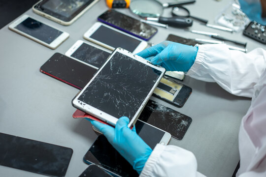 Forensic technician in blue gloves examines a cracked tablet among damaged smart phones and electronic devices on a laboratory table.