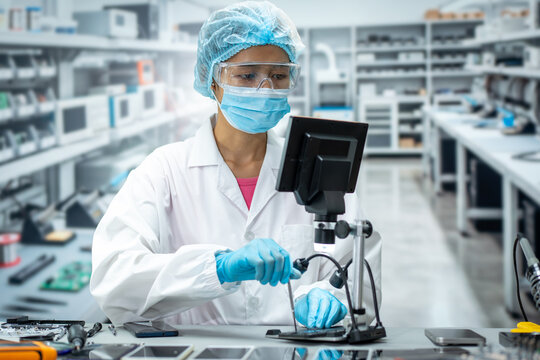 Female laboratory technician in protective clothing uses a digital microscope to inspect electronic parts in a clean modern research lab.