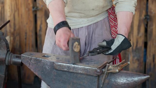 Adult blacksmith strikes metal on anvil with hammer while gloved craftsman steadies workpiece with tongs. Focused metalworker practices traditional blacksmithing in rustic workshop.