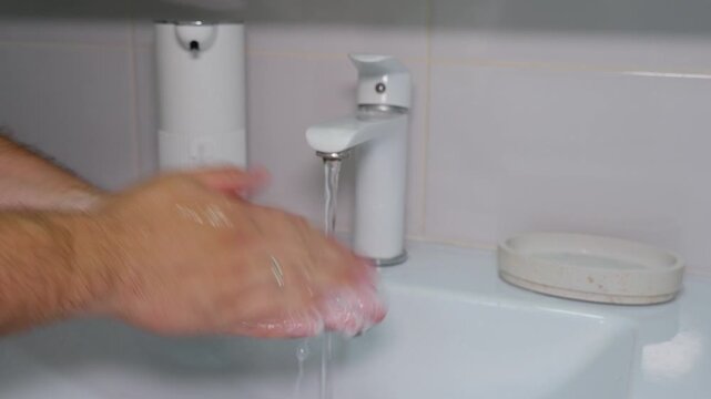 Man washes hands at sink under running water with concentrated expression, doing careful hygiene routine. Adult male rubs wet hands together in clean bathroom with white faucet and soap dish.