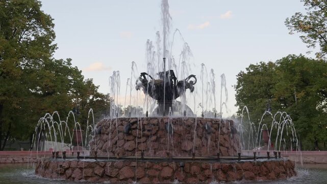 Ornamental fountain sends water upward around dark sculptural centerpiece in tree lined park. Stone fountain displays layered jets and flowing basin water in peaceful daylight, with arcing streams.