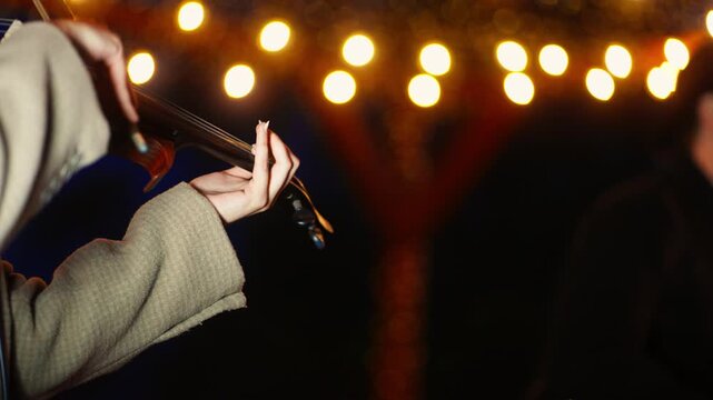 Close-up nighttime scene of a violinist performing under warm string lights, highlighting expressive hand movements, textured coat details, and atmospheric bokeh glow