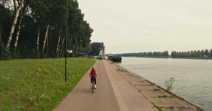 Drone tracking shot of a woman leisurely cycling along a scenic cycle path beside a river lined with trees, surrounded by lush green nature and peaceful countryside.