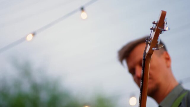 Close-up of musician playing electric guitar outdoors at dusk, warm string lights glowing in soft bokeh behind performer hands during intimate live performance